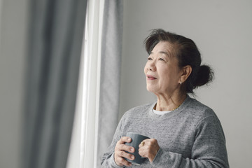 Asian senior woman drinking hot tea near window outdoor, lonely concept.