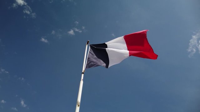 French flag, seen from below, flaps in the wind from a pole with blue sky behind it.