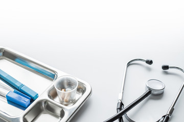 A stethoscope and a metal tray with thermometers and pills on white background, medical concept.