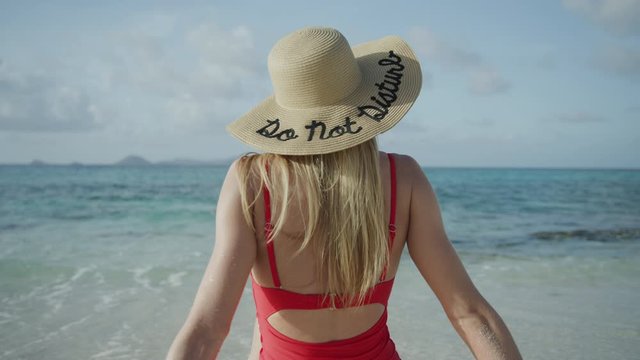 Rear view of woman sitting on ocean beach wearing do not disturb hat / Petit Tabac, Tobago Cays, St. Vincent and the Grenadines