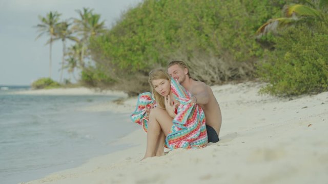 Slow motion of man bringing warm blanket to woman sitting on ocean beach / Petit Tabac, Tobago Cays, St. Vincent and the Grenadines