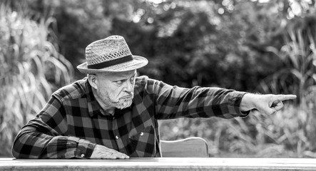 An elderly man sits with an angry face at a table in the park. He points to something with his...