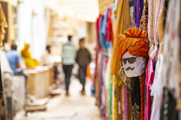 Fototapeta na wymiar (Selective focus) Beautiful shop with colorful traditional Indian clothes (Sari) and a mask depicting a person's face from Rajasthan. Jaisalmer, India.