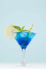 Summer cold blue lagoon drink with ice cubes, lemon slice, yellow straw, green mint in elegant wine glass on pastel mint background, vertical.