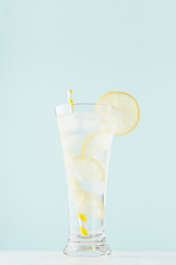 Detox cold tonic water with sunny lemon slices, ice cubes, yellow striped straw in elegant glass on white wood table, mint color wall, vertical.