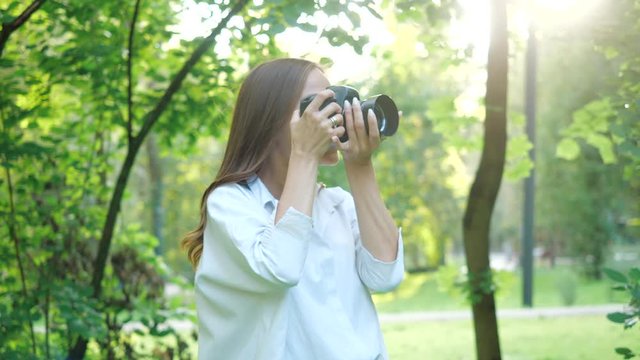 Pretty smiling girl photographer wearing white shirt is making photos with professional camera in a park on a soft background of green foliage and spraying water.