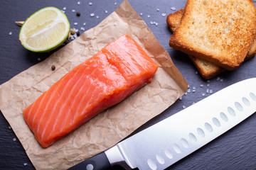 Salmon, avocado, lime and toasts laying with knife on slate background.