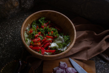 Cooking vegetable salad with lots of green, tomatoes, lettuce, and red onion. Wooden bowl with vegetable ingredients and wooden desk with cut red onion on it. Organic cooking series photography