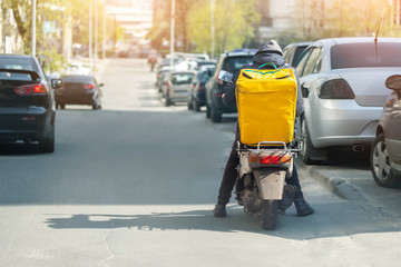 Food delivery courier with big yellow backpack riding scooter on city street with traffic. Fast lunch takeaway delivery. Teenager job