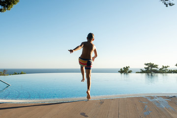 Boy jumping in swimming pool at sunset. European boy in colorful swimming trunks is running to jump into swimming pool during sunset with sea line on the background. Jumping set