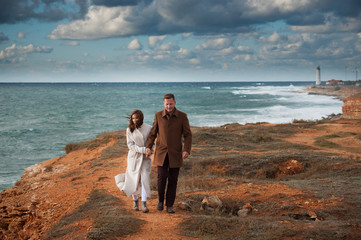 beautiful young couple bearded man in coat holding his woman with hair blown with wind walking along brown sea coast with lighthouse and moody sky with clouds on horizon