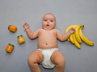Surprised baby on a gray background with bananas and jars of baby food.