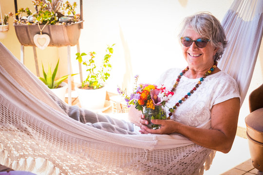 Relaxed senior woman lying down in a white hammock holding vase of flowers.  Grey hair and sunglasses. Serene people sitting in the terrace. Wooden table