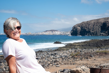 Portrait of attractive senior woman gray hair sitting on the beach in casual clothing. Horizon on the water. Blue sky and sea in the background. Holidays or pension. Serenity and relaxation