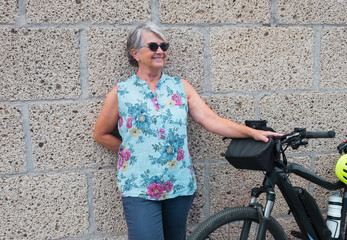 Fototapeta na wymiar Attractive senior woman people standing against a wall resting after an excursion with the e bike. Smiling. Retired woman casual clothing. Yellow helmet. Healthy lifestyle