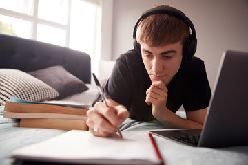 Male College Student Wearing Headphones Lies On Bed In Shared House Working On Laptop