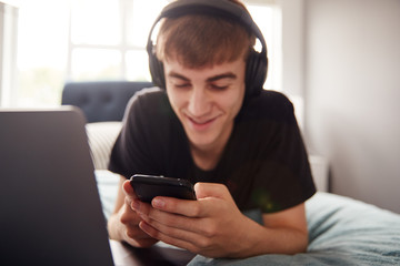 Male College Student Wearing Headphones Lies On Bed In Shared House With Mobile Phone Using Laptop