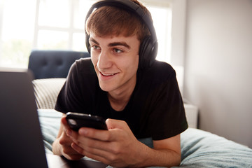 Male College Student Wearing Headphones Lies On Bed In Shared House With Mobile Phone Using Laptop