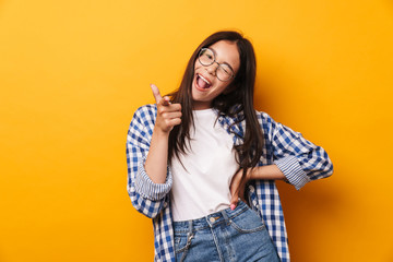 Smiling emotional young cute teenage girl in glasses posing isolated over yellow wall background pointing to you.