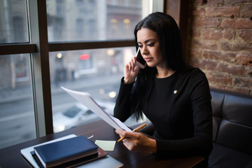 Young caucasian woman prosperous business worker holding paper document with monthly revenue report and talking with partner via cell telephone while sitting in restaurant. Manager reading resume - obrazy, fototapety, plakaty