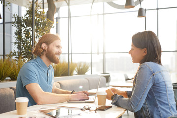Obraz premium Young bearded businessman talking to young businesswoman while working on laptop computer at the table during meeting in cafe
