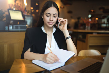 Young woman smart university student writing information in textbook during mobile phone conversation while sitting in modern restaurant. Female successful  manager talking via cellphone