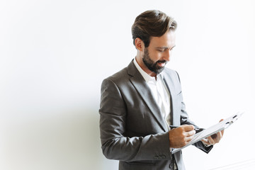 Image of satisfied successful businessman holding clipboard and writing down notes while working in office