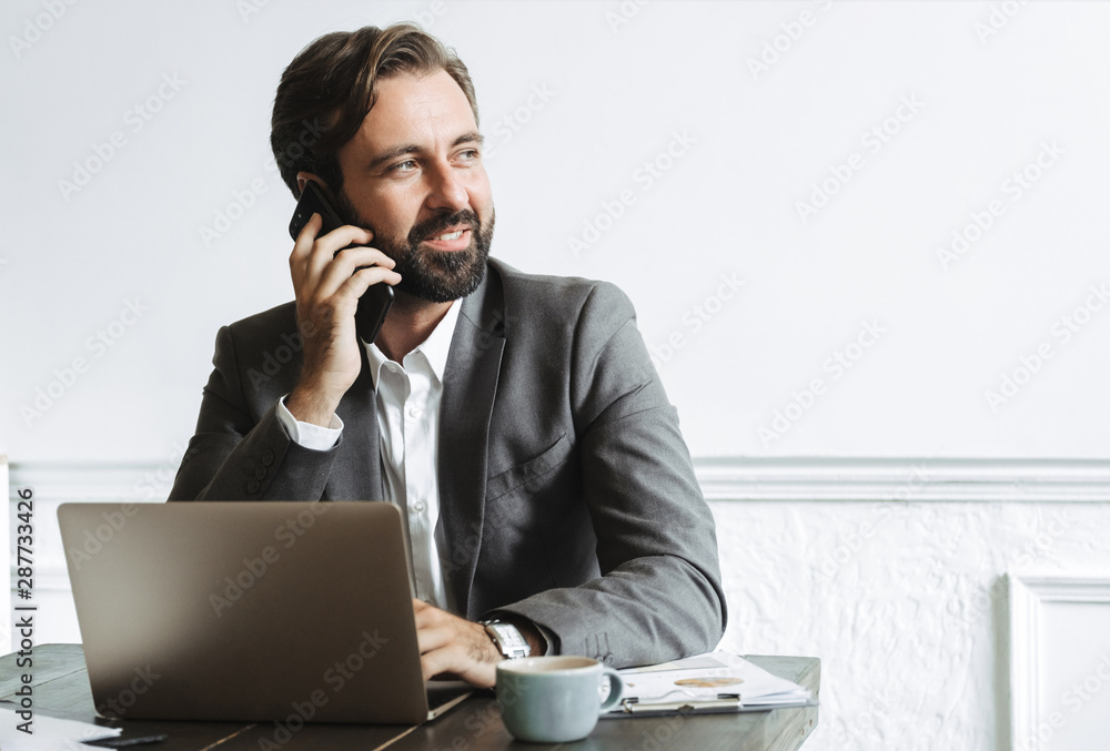 Sticker Image of smiling confident businessman typing on laptop and talking on cellphone while working in office