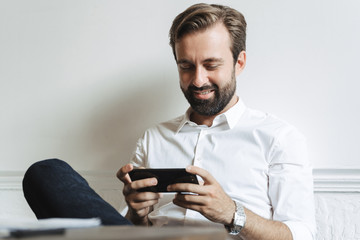 Image of successful smiling businessman playing video game on cellphone while sitting at table in office