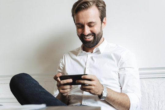 Image of successful laughing businessman playing video game on cellphone while sitting at table in office