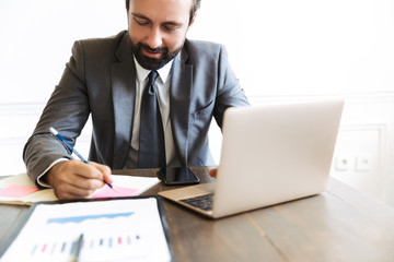 Image of concentrated confident businessman working at laptop and cellphone in office while writing down notes