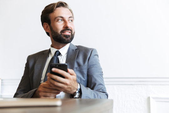 Image of bearded satisfied businessman typing on cellphone and looking aside while working in office