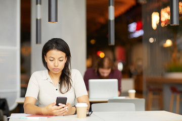 Serious young woman sitting at the table and using mobile phone for online work with man in the background in cafe