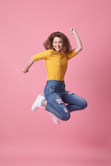Portrait of a joyful young woman in yellow shirt jumping and celebrating over pink background.