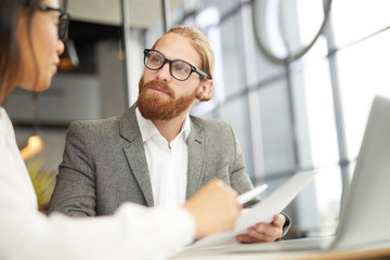 Serious bearded businessman in eyeglasses holding document and listening to his partner during business meeting at office