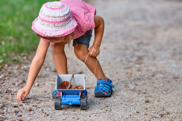 A child in summer clothing on a gravel footpath picking up something from the ground to put it into...