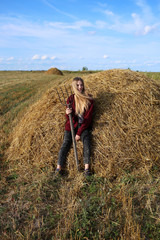 Naklejka na ściany i meble A cheerful, attractive girl farmer sits on the hay with a pitchfork in her hands and looks away smiling.