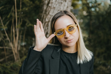 Blonde girl dressed in black jacket with yellow glasses with false eyelashes. Beauty, fashion concept. Pure skin. Jungle forest background. Classic suit style.