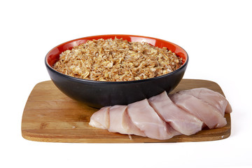 Sprouted wheat porridge in red and black bowl with sliced raw chicken meat near it. On wooden desk. Isolated on white background. Idea for healthy diet meal.