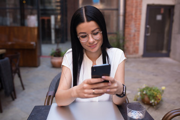 Fashionable female skilled lifestyle blogger reading article on web site via mobile phone while sitting in coffee shop during recreation time. Woman student typing text on cell telephone