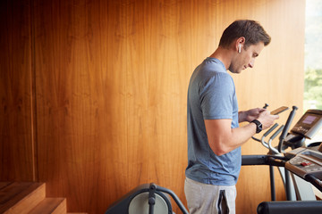 Fototapeta na wymiar Man Exercising On Treadmill At Home Wearing Wireless Earphones Checking Mobile Phone