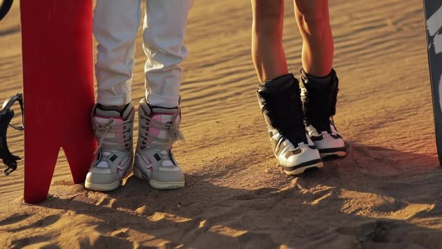 sandboard, sandboarding . feet closeup, two girls in boots with snowboard standing on the sand and preparing for the descent. sunset on desert