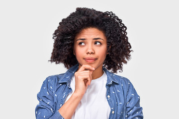 Doubtful African American woman has puzzled expression, frowns her face and looking at one side with hand on chin. Dark skinned female can`t make choice, has questioned expression, isolated on white.