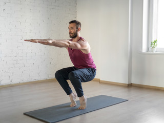 Chair pose. Caucasian fit man stands on tiptoe and doing yoga in fitness studio, selective focus.