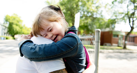 A portrait of trisomie 21 child girl outside hugging his mother on a school playground