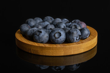 Lot of whole fresh blue bilberry on round bamboo coaster isolated on black glass