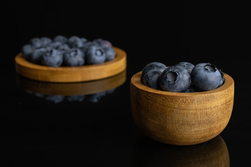 Lot of whole fresh blue bilberry in tiny wooden bowl on round bamboo coaster isolated on black glass