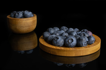 Lot of whole fresh blue bilberry in tiny wooden bowl on round bamboo coaster isolated on black glass