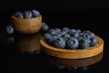 Lot of whole fresh blue bilberry in tiny wooden bowl on round bamboo coaster isolated on black glass