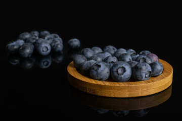 Lot of whole fresh blue bilberry on round bamboo coaster isolated on black glass